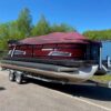 A maroon and black Starcraft pontoon boat with a matching maroon protective cover, sitting on a dual-axle LoadRite trailer parked on asphalt under a clear blue sky.