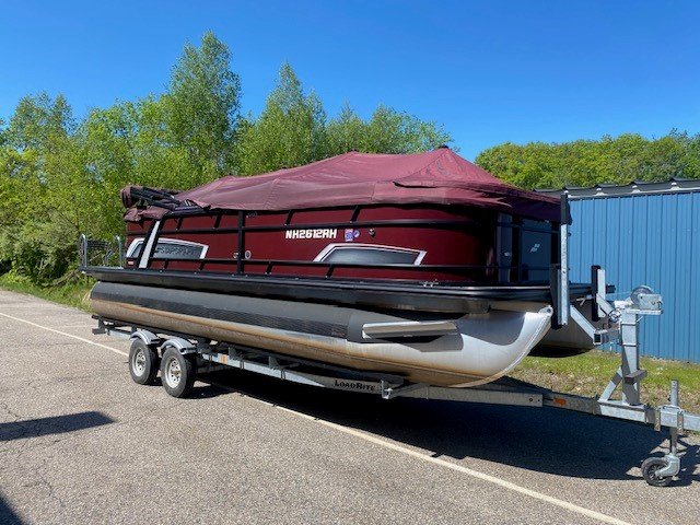 A maroon and black Starcraft pontoon boat with a matching maroon protective cover, sitting on a dual-axle LoadRite trailer parked on asphalt under a clear blue sky.
