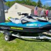 A teal and gray Sea-Doo personal watercraft with bright yellow lettering sitting on a single-axle Venture trailer parked on a grassy area next to a building.