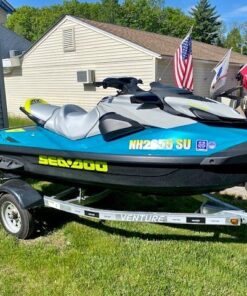 A teal and gray Sea-Doo personal watercraft with bright yellow lettering sitting on a single-axle Venture trailer parked on a grassy area next to a building.