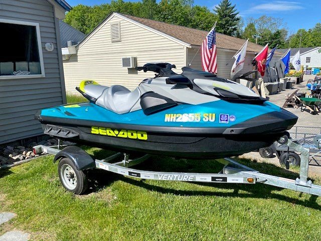 A teal and gray Sea-Doo personal watercraft with bright yellow lettering sitting on a single-axle Venture trailer parked on a grassy area next to a building.