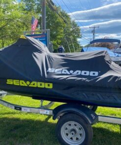 A teal and gray Sea-Doo personal watercraft with bright yellow lettering sitting on a single-axle Venture trailer parked on a grassy area next to a building.