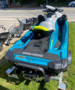 A teal and gray Sea-Doo personal watercraft with bright yellow lettering sitting on a single-axle Venture trailer parked on a grassy area next to a building.
