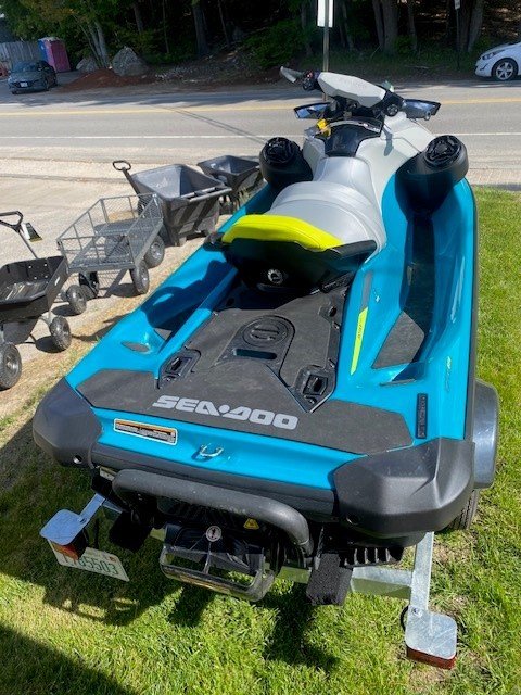 A teal and gray Sea-Doo personal watercraft with bright yellow lettering sitting on a single-axle Venture trailer parked on a grassy area next to a building.