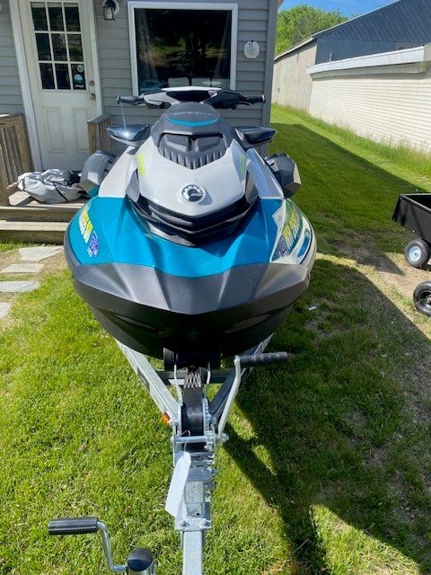 A teal and gray Sea-Doo personal watercraft with bright yellow lettering sitting on a single-axle Venture trailer parked on a grassy area next to a building.