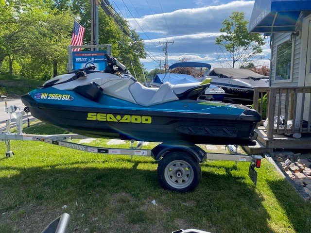 A teal and gray Sea-Doo personal watercraft with bright yellow lettering sitting on a single-axle Venture trailer parked on a grassy area next to a building.